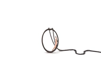 Puede incluir: Un collar de plata oxidada negra con un colgante de oro rosa. El colgante es un círculo con un círculo más pequeño cortado. La cadena es delicada y tiene un acabado oxidado negro.