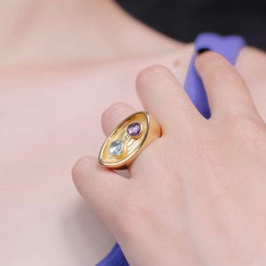 A woman wears a striking gold plated ring adorned with a purple stone and a blue gem, reflecting the ring's artistic and elegant design.