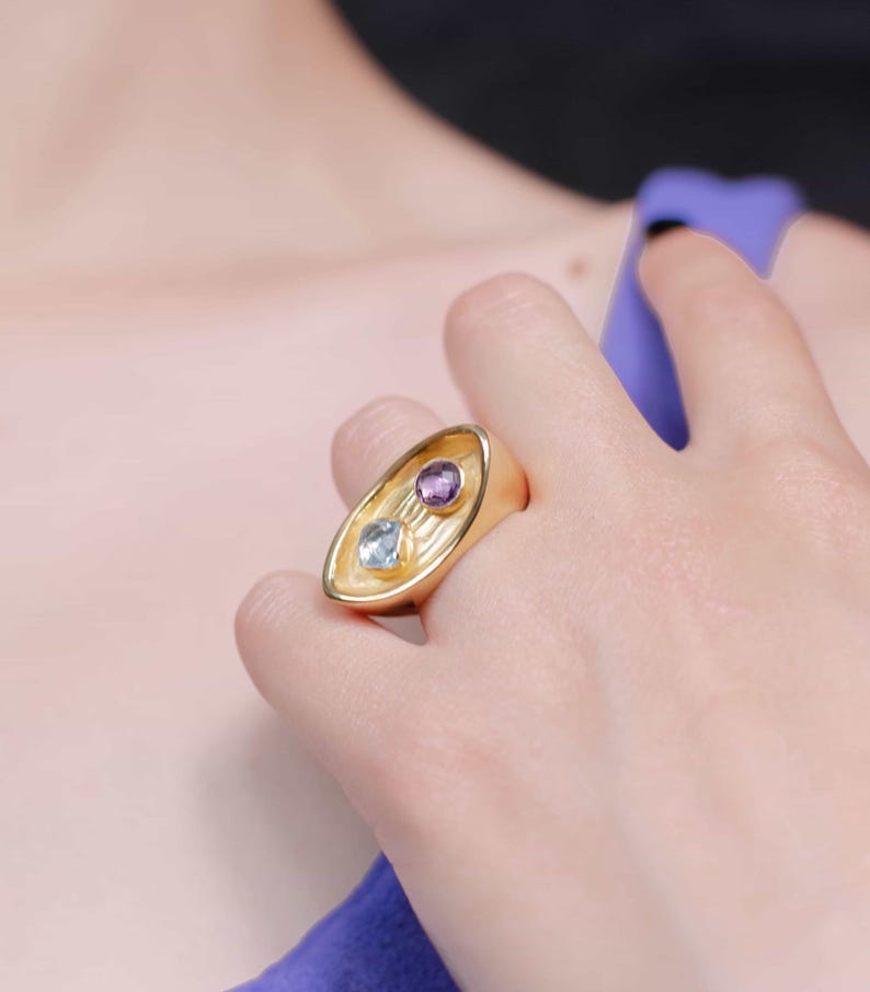 A woman wears a striking gold plated ring adorned with a purple stone and a blue gem, reflecting the ring's artistic and elegant design.