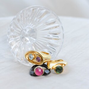 A book, a bold two-stone gold ring featuring amethyst and topaz, and a glass of wine, representing style and sophistication.