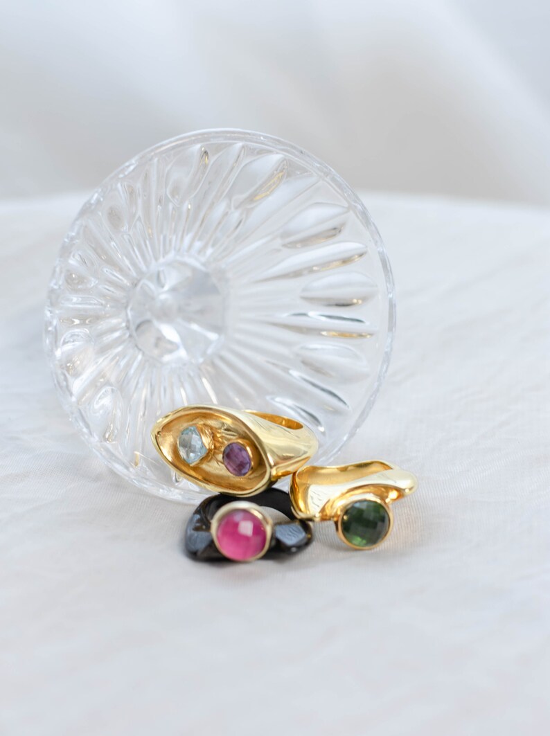 A book, a bold two-stone gold ring featuring amethyst and topaz, and a glass of wine, representing style and sophistication.