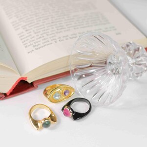 A book, a chunky gold-plated ring with amethyst and topaz, and a glass of wine, symbolizing elegance and creativity.