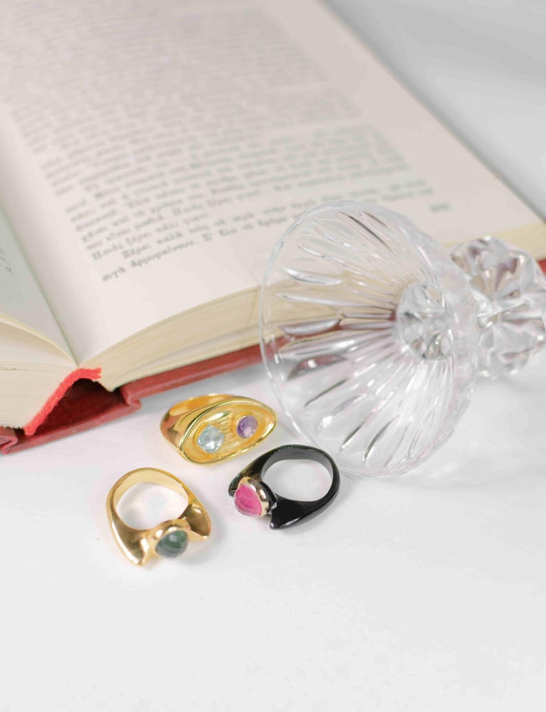A book, a chunky gold-plated ring with amethyst and topaz, and a glass of wine, symbolizing elegance and creativity.