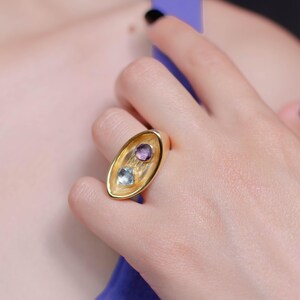 A woman showcases a chunky gold plated two stone ring featuring a purple amethyst and a blue topaz, highlighting its unique design and elegance.