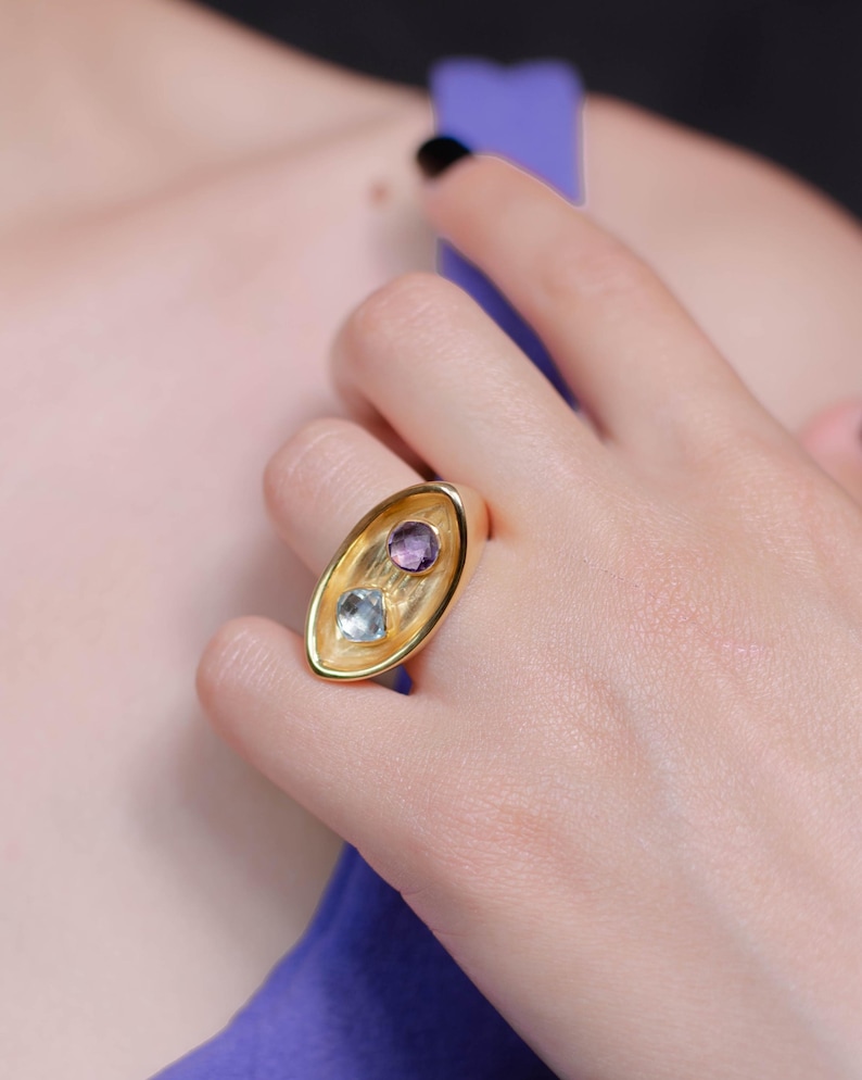A woman showcases a chunky gold plated two stone ring featuring a purple amethyst and a blue topaz, highlighting its unique design and elegance.