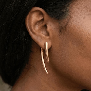 Peut inclure: Gros plan sur une oreille portant des boucles d'oreilles dorées. Les boucles d'oreilles ont un design unique, avec une forme incurvée et allongée qui pend du lobe de l'oreille. Les boucles d'oreilles ont une finition polie, reflétant la lumière.