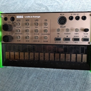 Może przedstawiać: Czarno-złoty syntezator KORG volca keys w zielonej ochronnej obudowie. Syntezator posiada rząd klawiszy, pokręteł i przycisków do sterowania parametrami dźwięku. Tekst "KORG volca keys" jest nadrukowany na przednim panelu.