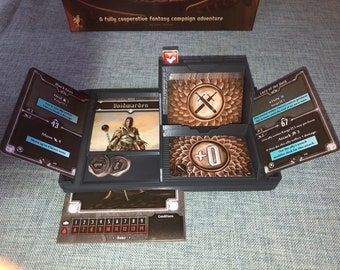 May include: A close-up of a board game with a black, brown, and wooden game board. The game board has a card slot with a card that reads "Voidwarden". There are two coins on the board. The game board also has a card slot with a card that reads "Lure of the Wild". The card has a blue background and the text "STUN" and "Attack 3" is visible. The game board also has a card slot with a card that reads "+0".
