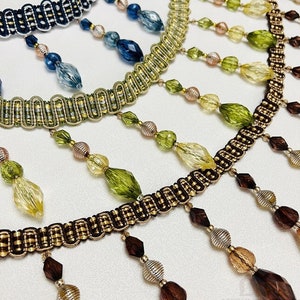 Op de afbeelding: Drie strengen met kralenversiering met kralen in verschillende kleuren. De bovenste streng heeft blauwe kralen, de middelste streng heeft groene kralen en de onderste streng heeft bruine kralen. Elke streng heeft een gouden en zwarte geweven versiering.