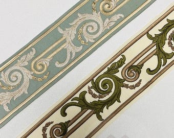 Pode incluir: Duas tiras de fita decorativa de tecido com um padrão bordado de volutas de folhas de acanto. Uma tira é azul-clara com bordado creme, e a outra é creme com bordado verde-oliva. Ambas apresentam linhas paralelas e um logotipo.
