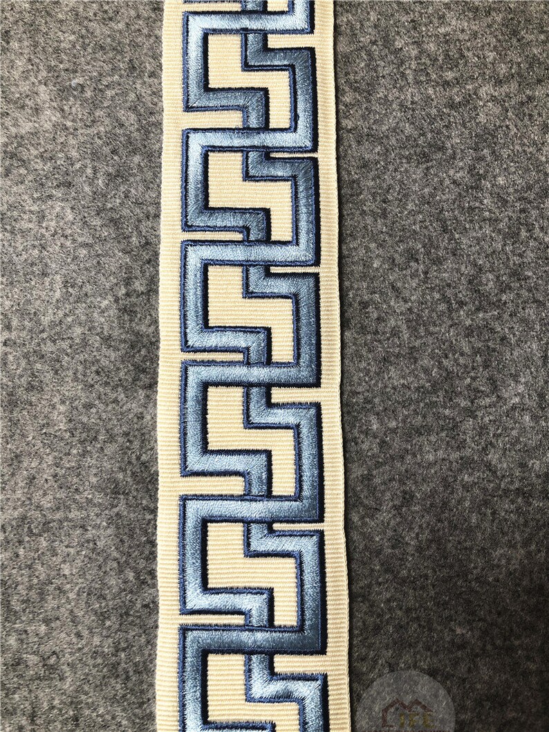 May include: A white ribbon with a blue geometric pattern. The pattern is a repeating design of interlocking squares and rectangles.