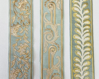 Pode incluir: Três fitas de tecido diferentes com fundo azul claro e bordados em dourado e branco. As fitas apresentam desenhos florais e de folhas.