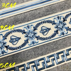 May include: Three different trims of embroidered ribbon in cream with blue accents. The trims feature a floral design, a bee design, and a Greek key design. The trims are labeled 3CM, 9CM, and 5CM.