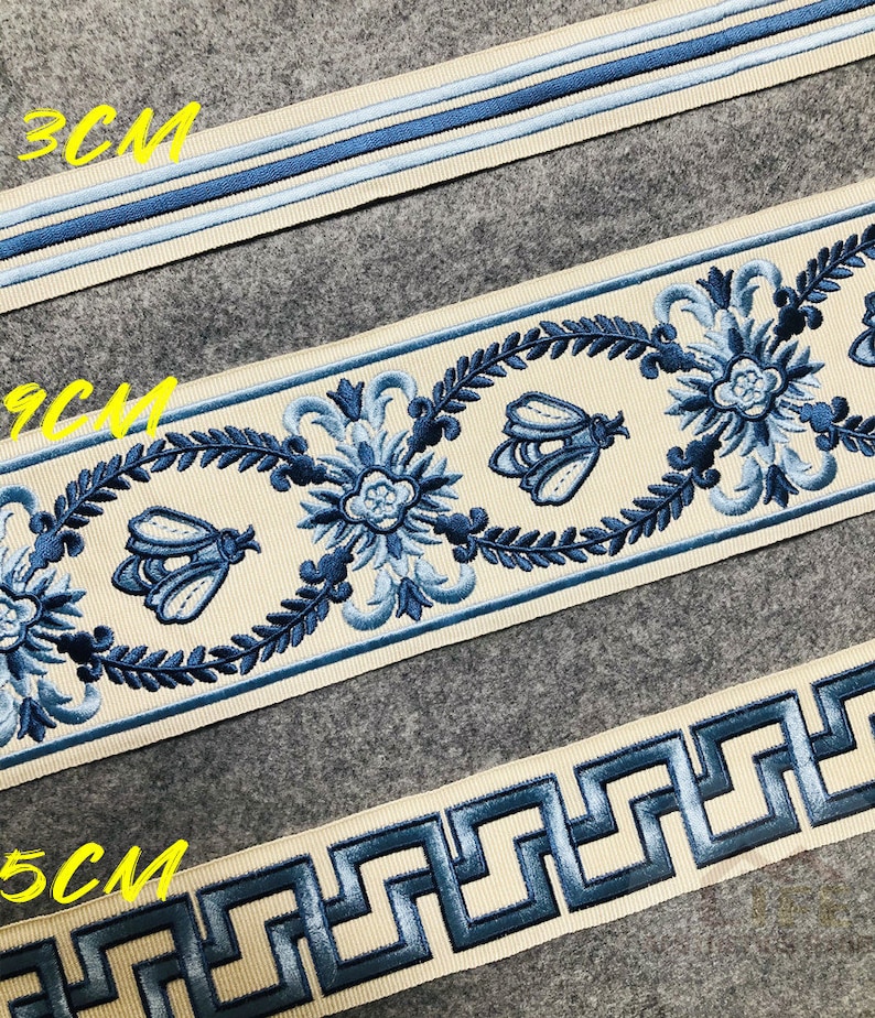 May include: Three different trims of embroidered ribbon in cream with blue accents. The trims feature a floral design, a bee design, and a Greek key design. The trims are labeled 3CM, 9CM, and 5CM.