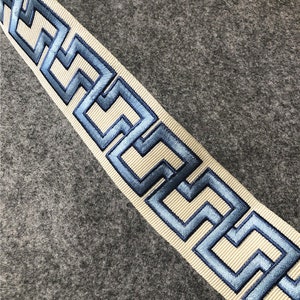 May include: A white ribbon with a blue embroidered Greek key pattern. The ribbon is on a gray background.