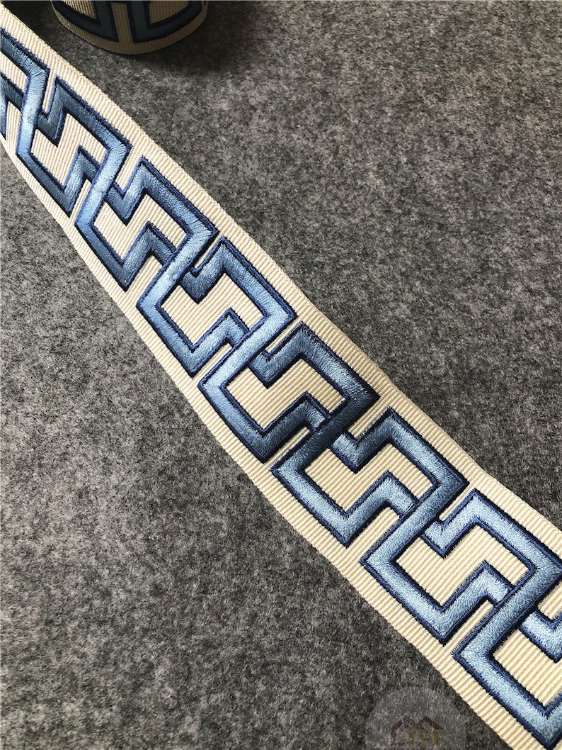 May include: A white ribbon with a blue embroidered Greek key pattern. The ribbon is on a gray background.