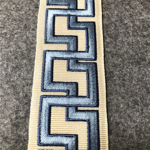 May include: A beige fabric trim with a blue embroidered geometric pattern. The pattern is a repeating design of interlocking squares.