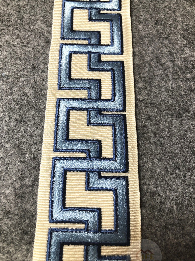 May include: A beige fabric trim with a blue embroidered geometric pattern. The pattern is a repeating design of interlocking squares.