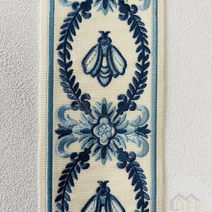 May include: A white ribbon with a blue floral and bee design. The ribbon has a repeating pattern of a stylized flower with a bee in the center, surrounded by leaves.