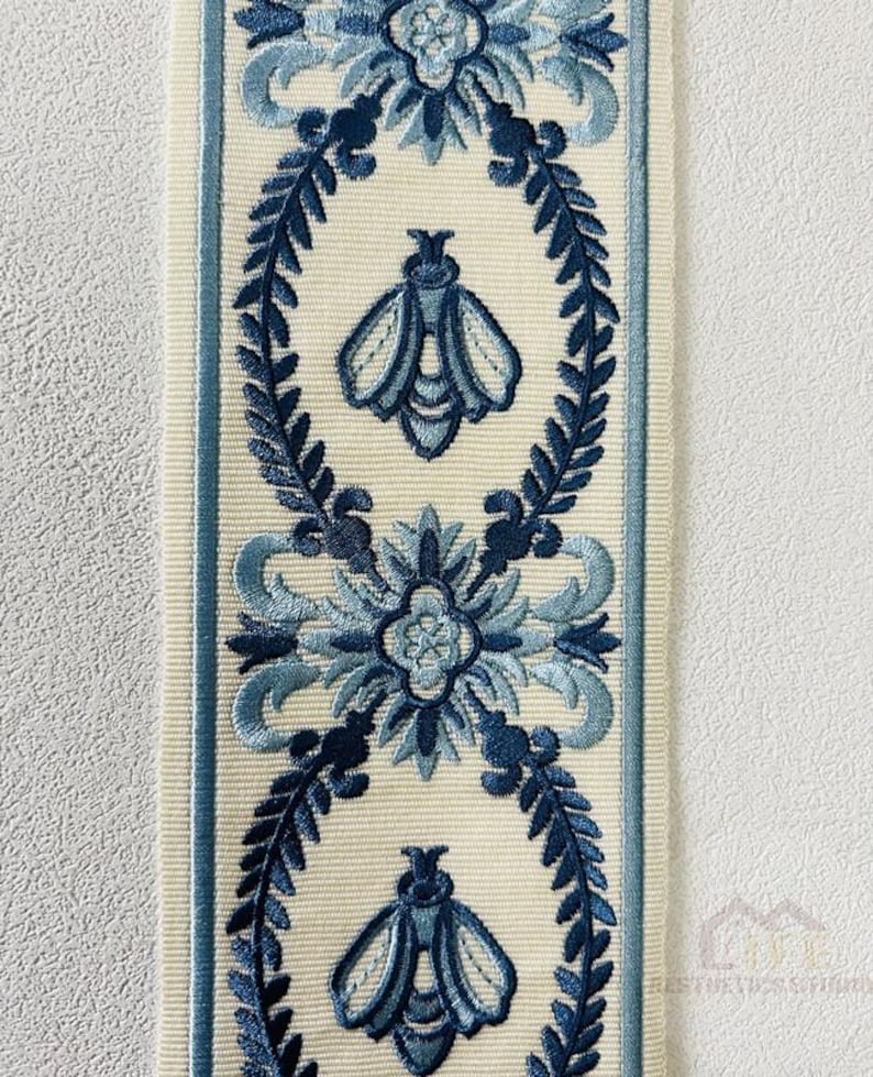 May include: A white ribbon with a blue floral and bee design. The ribbon has a repeating pattern of a stylized flower with a bee in the center, surrounded by leaves.