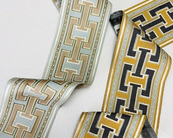 May include: Decorative ribbon in two color schemes. One ribbon features a light blue, tan, and cream geometric pattern. The other ribbon has a mustard yellow, black, and cream geometric design. Both ribbons have a repeating chain-like border.