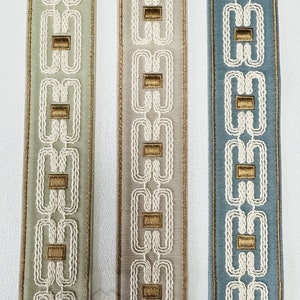 May include: Close-up of three decorative ribbons in shades of green, gray, and blue. Each ribbon features a repeating pattern of white interlocking shapes with gold-colored rectangular accents and gold trim along the edges. The ribbons are vertically aligned.