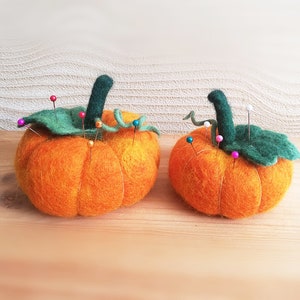 Puede incluir: Dos calabazas de fieltro naranja con hojas verdes y alfileres de colores. Las calabazas son una forma divertida y decorativa de sujetar alfileres y agujas.