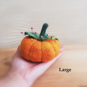 Puede incluir: Un gran coj&iacute;n de alfileres de fieltro naranja en forma de calabaza con hojas de fieltro verde y un tallo. El coj&iacute;n est&aacute; lleno de alfileres de colores.