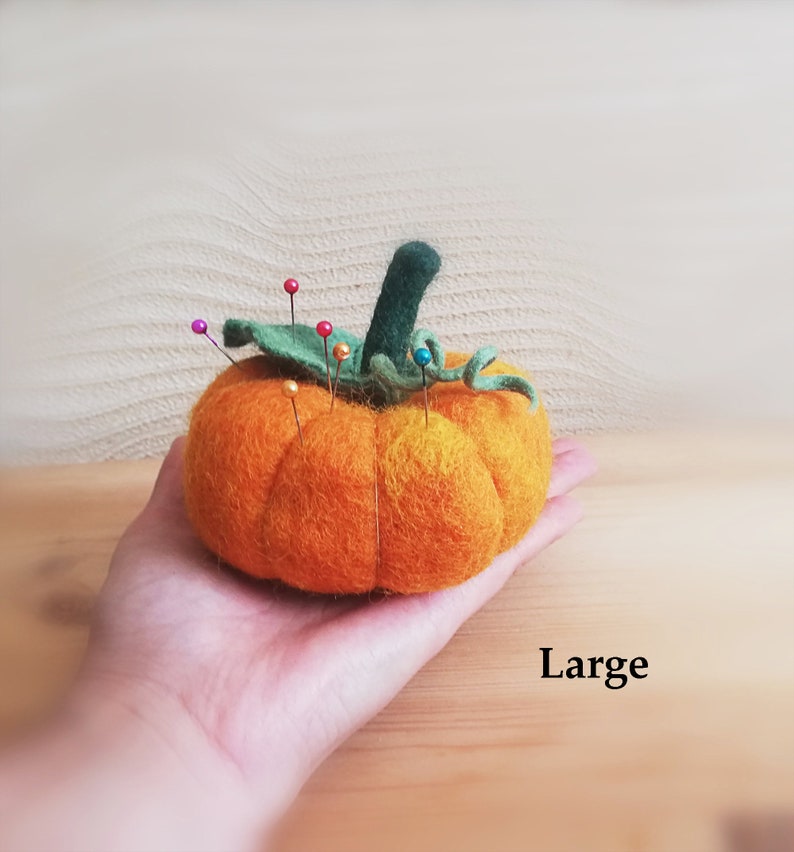 Puede incluir: Un gran coj&iacute;n de alfileres de fieltro naranja en forma de calabaza con hojas de fieltro verde y un tallo. El coj&iacute;n est&aacute; lleno de alfileres de colores.