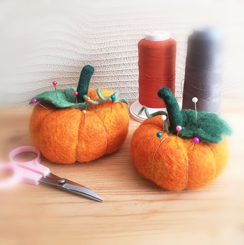 Puede incluir: Dos calabazas de fieltro naranja con hojas y tallos verdes, utilizadas como alfileteros. Las calabazas est&aacute;n llenas de alfileres de colores.