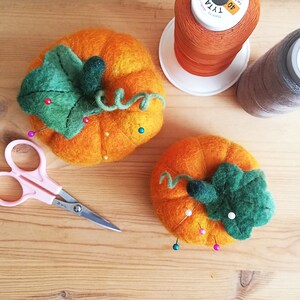 Puede incluir: Dos alfileteros de calabaza de fieltro con hojas y tallos verdes. Las calabazas son de color naranja y tienen alfileres clavados en ellas.