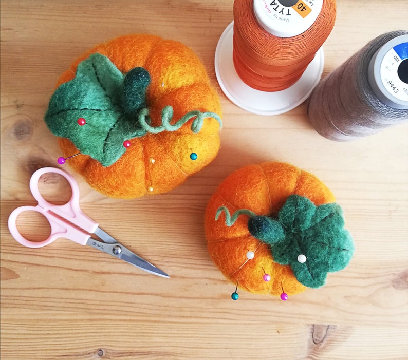Puede incluir: Dos alfileteros de calabaza de fieltro con hojas y tallos verdes. Las calabazas son de color naranja y tienen alfileres clavados en ellas.