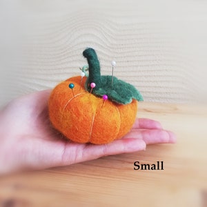 Puede incluir: Un peque&ntilde;o coj&iacute;n de alfileres de fieltro naranja en forma de calabaza con un tallo y hojas de fieltro verde. El coj&iacute;n est&aacute; lleno de alfileres con cabezas de colores.