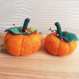 Puede incluir: Dos calabazas de fieltro naranja con tallos y hojas de fieltro verde. Las calabazas est&aacute;n llenas de alfileres y se utilizan como alfileteros.