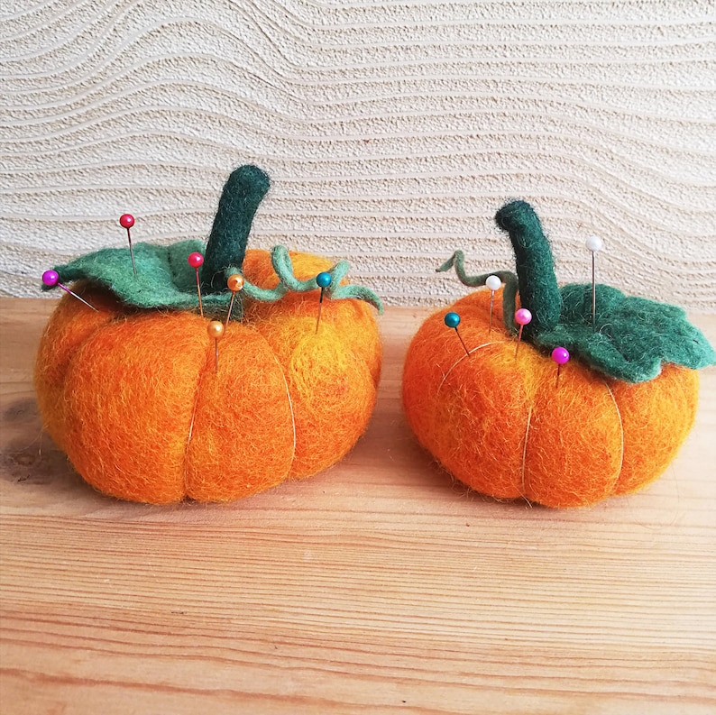 Puede incluir: Dos calabazas de fieltro naranja con tallos y hojas de fieltro verde. Las calabazas est&aacute;n llenas de alfileres y se utilizan como alfileteros.