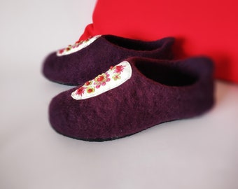 May include: A pair of purple wool slippers with a white embroidered floral design. The slippers have a rounded toe and a flat sole.