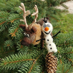 May include: A brown and white reindeer figurine with antlers and a white snowman figurine with black eyes and a carrot nose. Both figurines are made of felt and are sitting on a green pine branch.