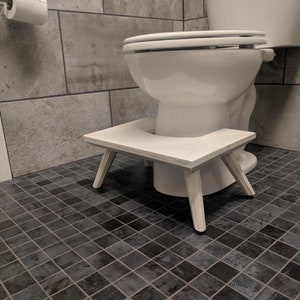 May include: White wooden toilet foot stool with a curved top and four legs. The stool is positioned in front of a white toilet in a bathroom with gray tile.