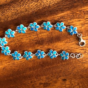 May include: A silver bracelet featuring a floral design with blue opal-like stones. Each flower is connected by small silver links, and the bracelet has a lobster clasp closure. The bracelet is displayed on a wooden surface.