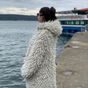 May include: A woman wearing a white fluffy hooded cardigan stands on a pier overlooking the water. The cardigan has a loose fit and a textured, fuzzy appearance.