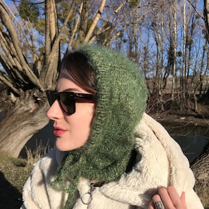 May include: A woman wearing a green knitted hat and a white faux fur jacket. She is wearing sunglasses and has her hair pulled back.