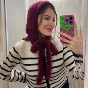May include: A woman wearing a burgundy fuzzy knit hat and matching scarf with a black and white striped sweater. She is holding a pink and green cell phone case with a dripping green design.