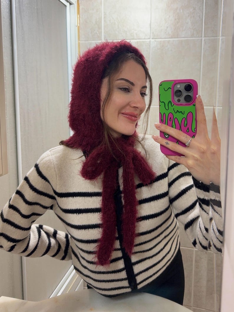 May include: A woman wearing a burgundy fuzzy knit hat and matching scarf with a black and white striped sweater. She is holding a pink and green cell phone case with a dripping green design.