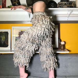 Puede incluir: Un pantalón beige con una textura única, peluda y con bucles. El pantalón está hecho de un material suave y peludo y tiene un corte holgado.