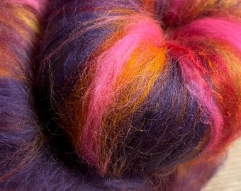 May include: Close-up of a vibrant, multicolored fiber art material. The material is a blend of pink, orange, and purple hues, creating a visually striking and textured appearance. The fibers are soft and fluffy, ideal for crafting projects.