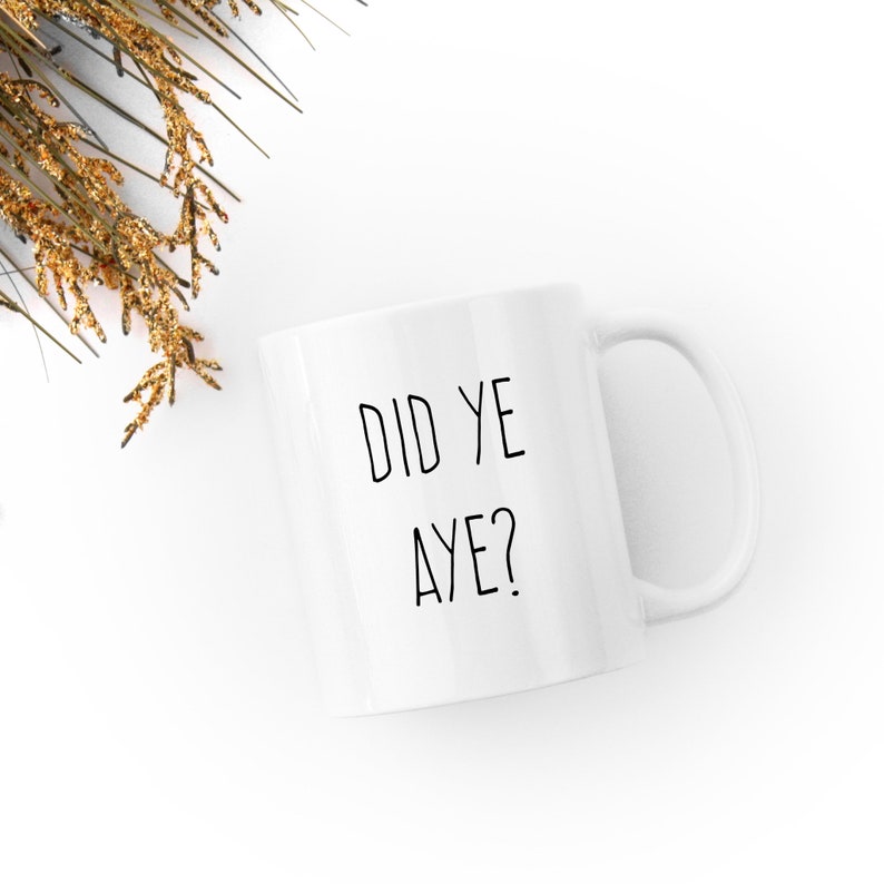 May include: White ceramic mug with the text "DID YE AYE?" in black. The mug has a curved handle and is set against a white background with a sprig of gold-colored decorative foliage.