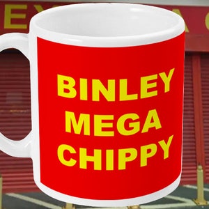 Puede incluir: Una taza de cerámica blanca con fondo rojo y texto amarillo que dice "Binley Mega Chippy". La taza está delante de un edificio rojo con un letrero amarillo que dice "Binley Chippy".