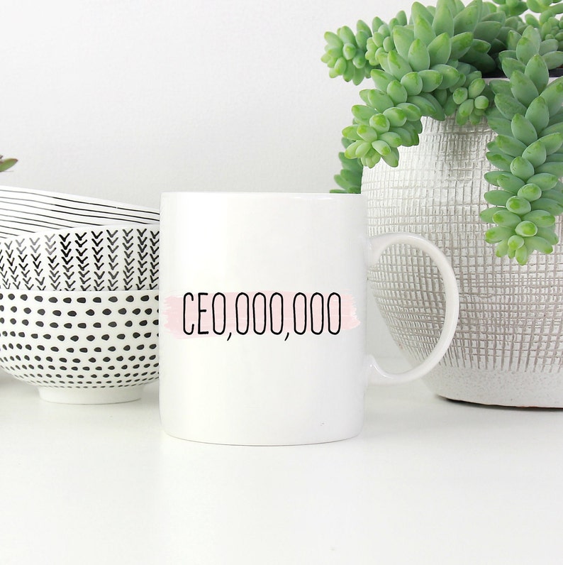 Op de afbeelding: Witte keramische mok met zwarte tekst die "CEO,000,000" luidt met een roze aquarelstreep achter de tekst.
