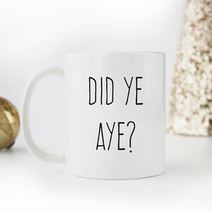 May include: White ceramic mug with the text "DID YE AYE?" in black, a humorous phrase. The mug has a curved handle and is set against a neutral background with a decorative gold ornament and a gold-colored tree.