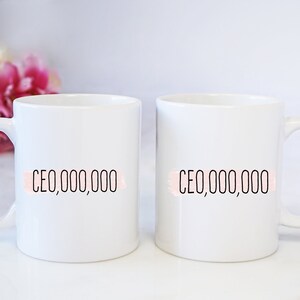 Op de afbeelding: Twee witte keramische mokken met zwarte tekst die "CEO,000,000" op elke mok staat. De tekst staat op een roze aquarel achtergrond.